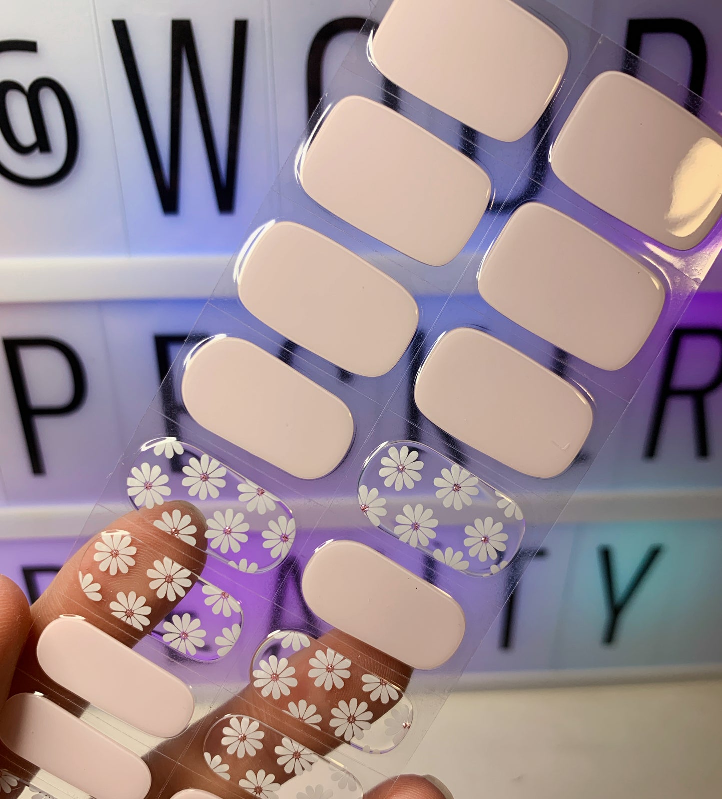 Hand holding a sheet of nail stickers with pink nude base and white daisy floral patterns.