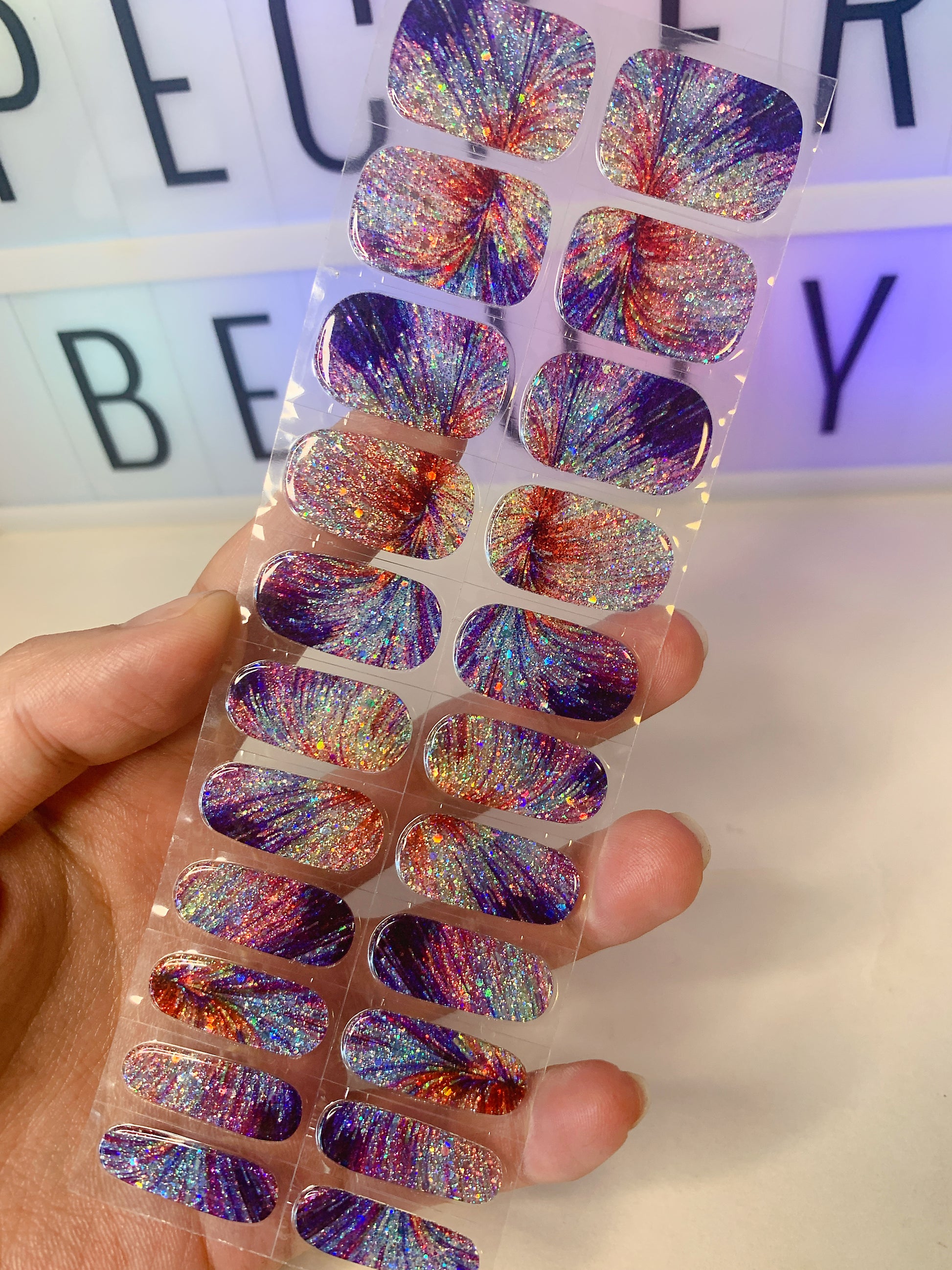 Hand holding nail wraps with colorful firework burst designs in purple, red, and gold glitter.
