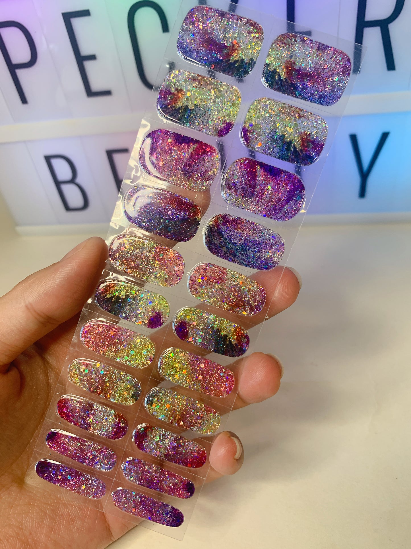 Hand holding a sheet of galaxy-themed glitter nail wraps with colorful sparkle.
