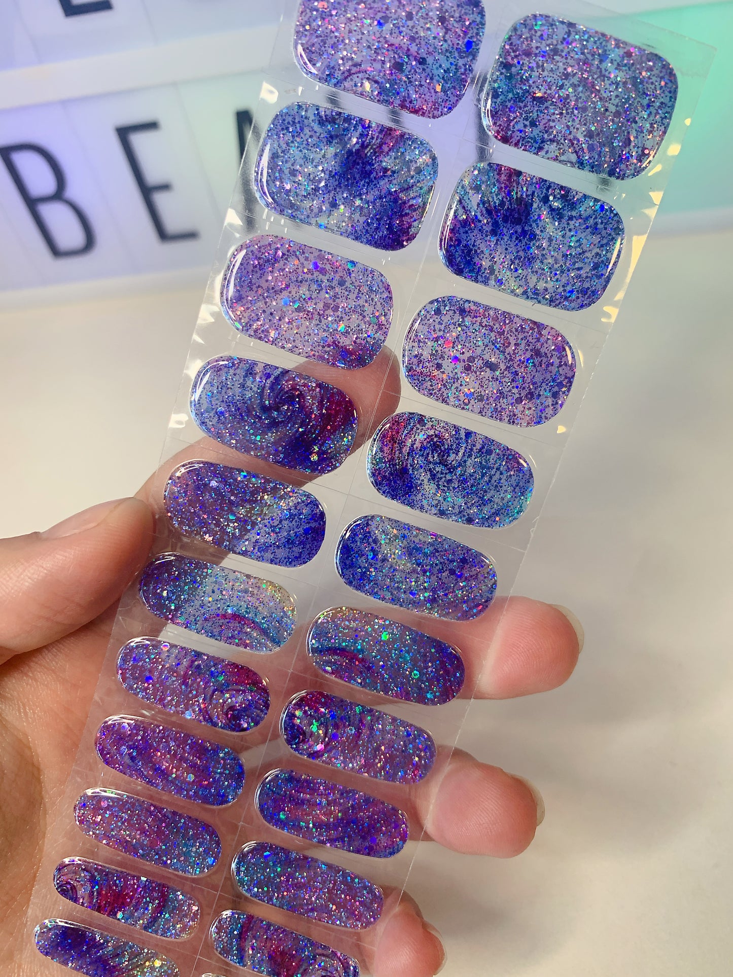 Hand holding a sheet of blue and purple galaxy glitter nail wraps with chunky sparkle.
