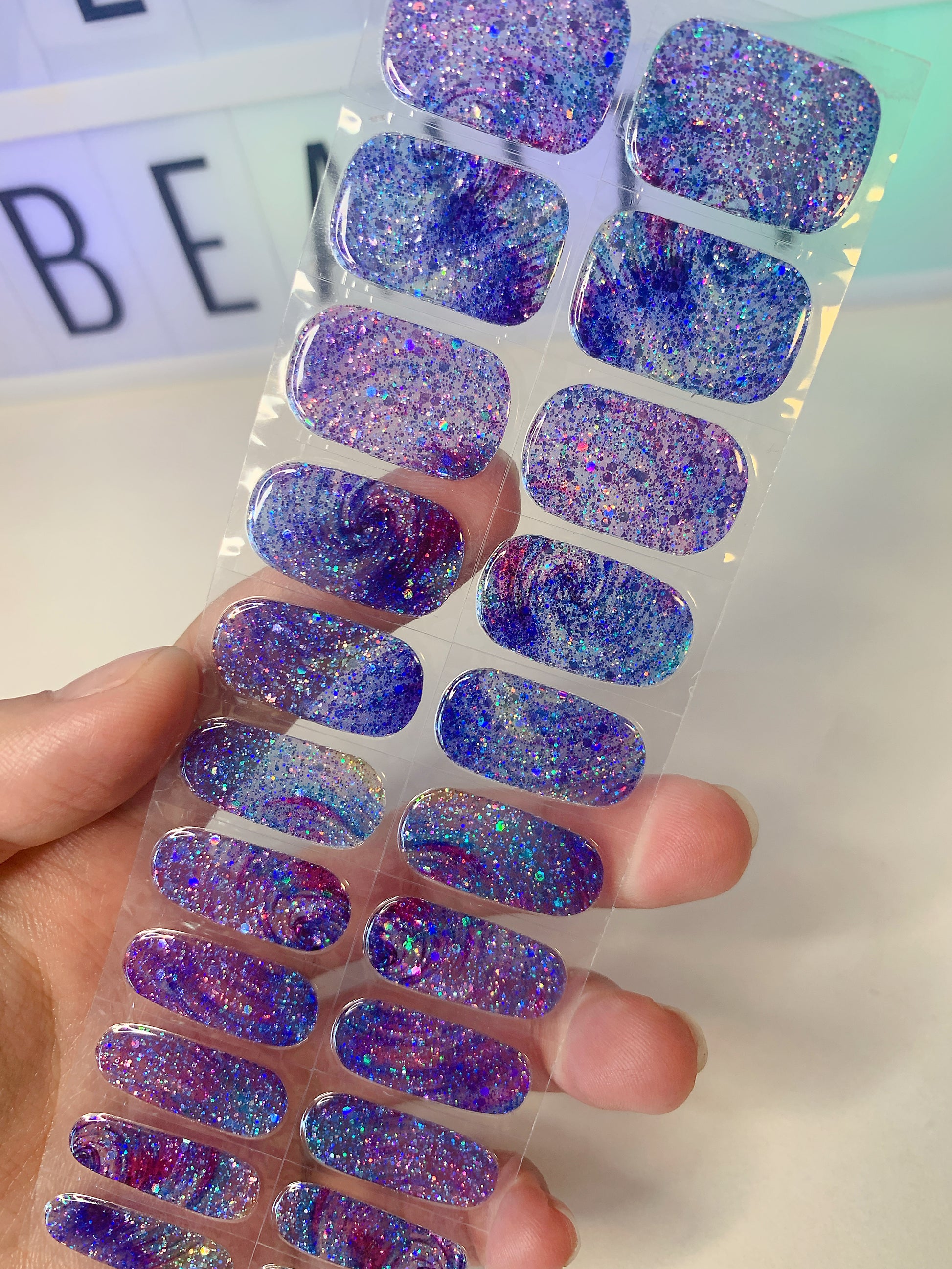 Hand holding a sheet of blue and purple galaxy glitter nail wraps with chunky sparkle.

