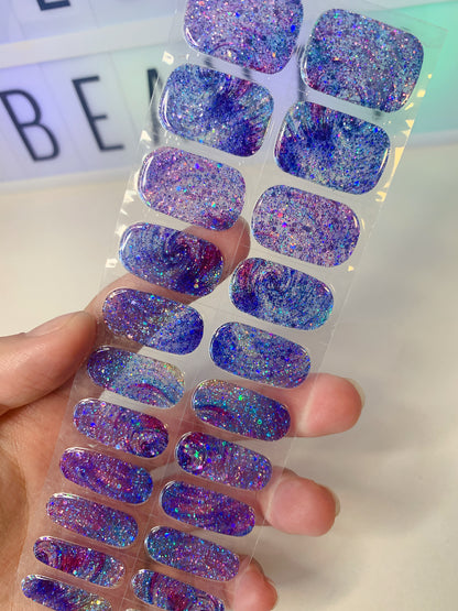 Hand holding a sheet of blue and purple galaxy glitter nail wraps with chunky sparkle.
