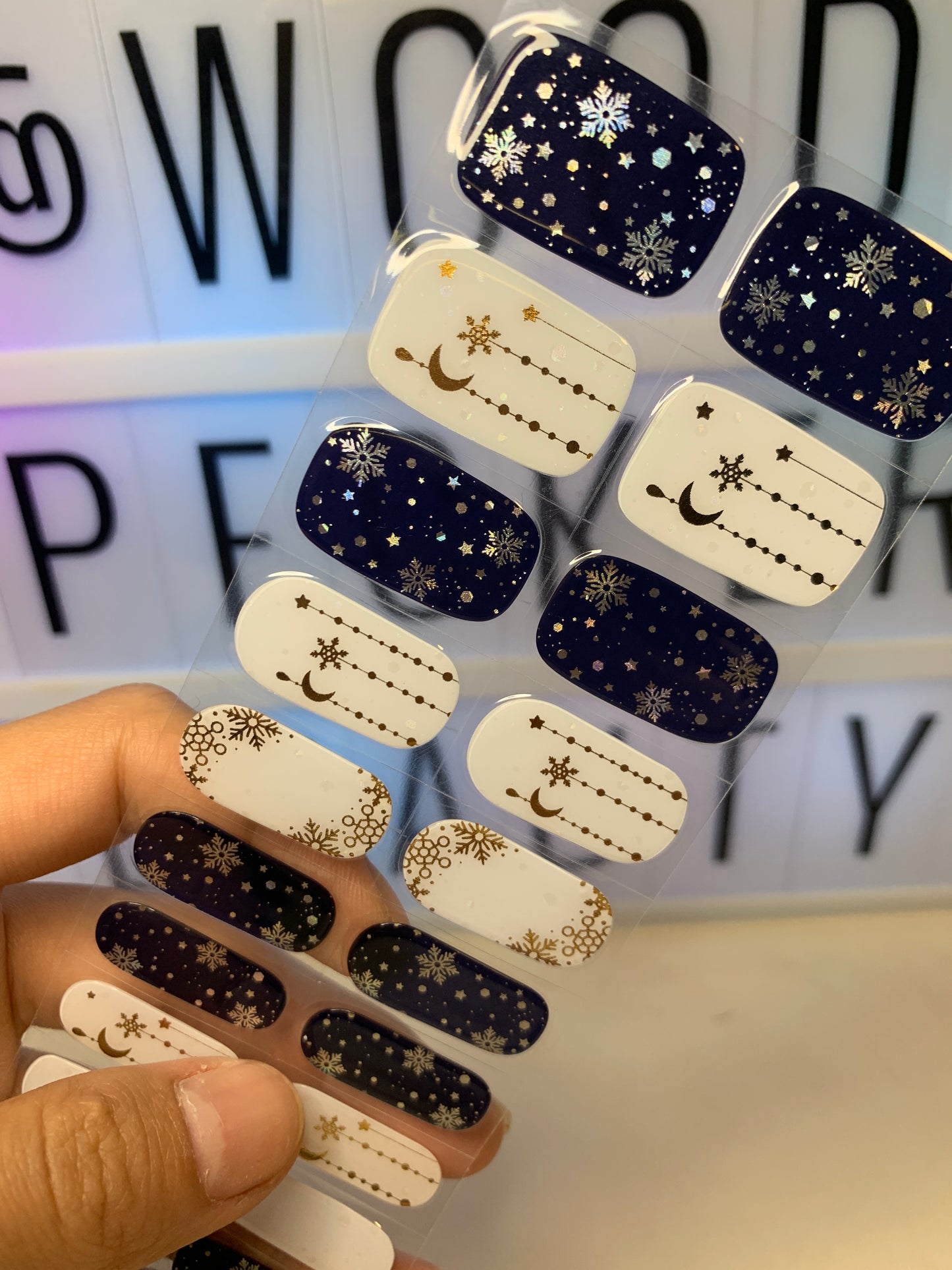 Hand holding a sheet of celestial nail stickers with starry night and snowflake patterns in navy and white.
