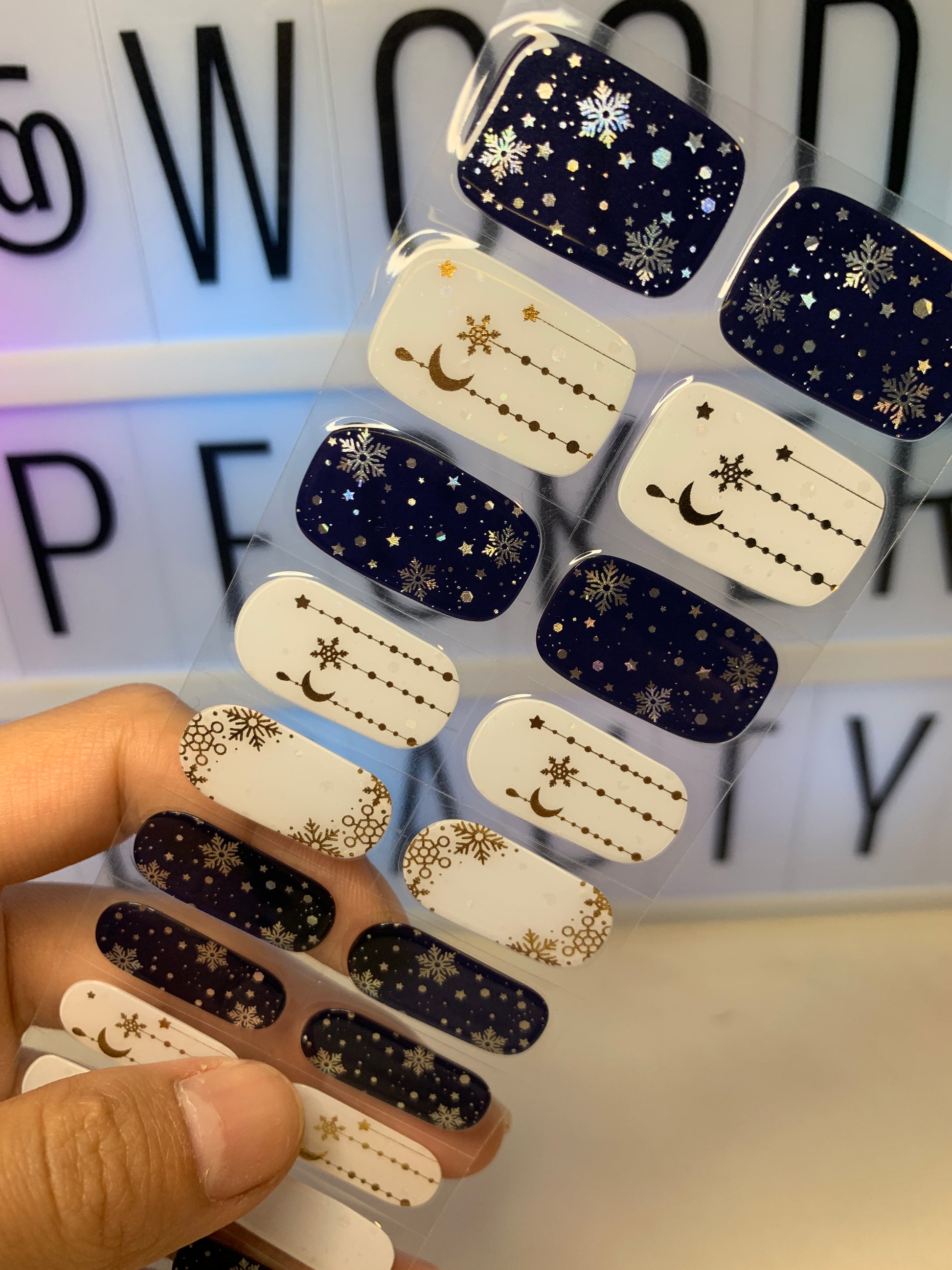 Hand holding a sheet of celestial nail stickers with starry night and snowflake patterns in navy and white.
