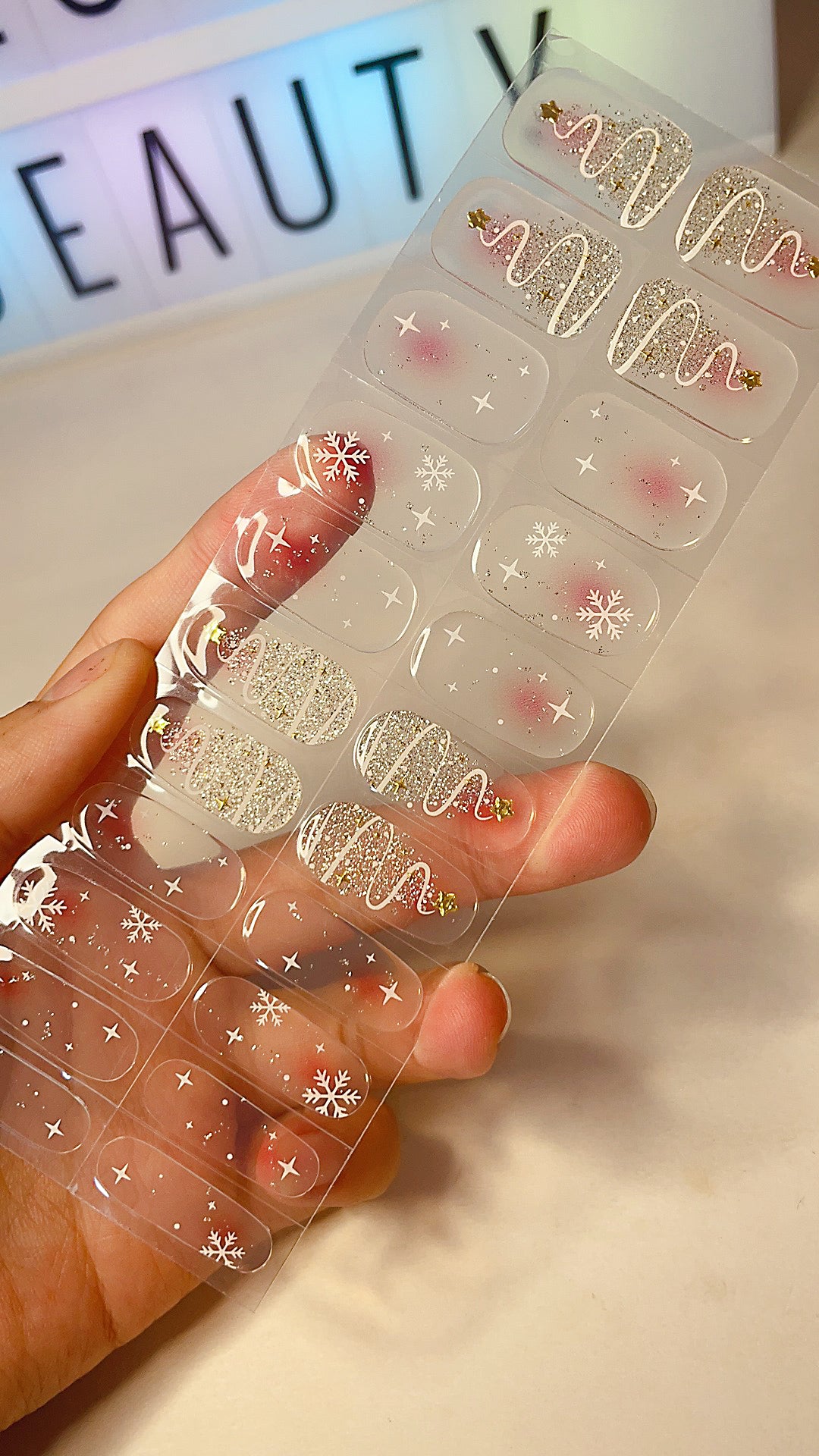 Hand holding a sheet of translucent pink Christmas nail wraps with gold glitter and snowflake accents.
