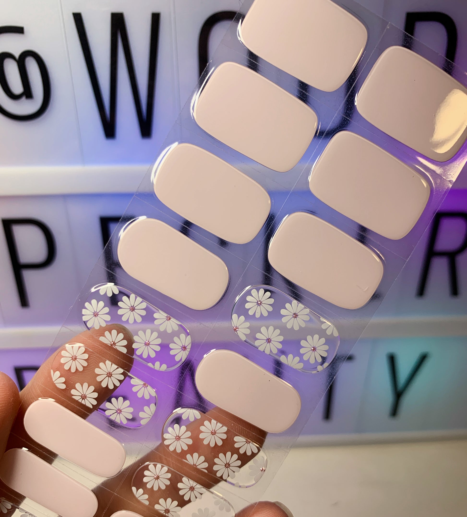 Hand holding a sheet of nail stickers with pink nude base and white daisy floral patterns.
