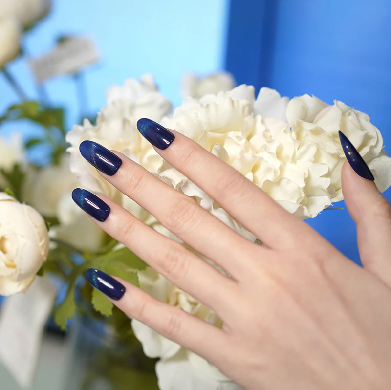 Hand with glossy navy blue nail stickers holding white flowers in a soft natural light setting.
